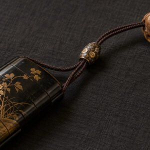 Illustration d’une perle ojime métallique à décor floral sur des cordons reliant un inro laqué et un netsuke en bois sculpté.