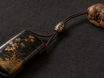 Ilustración de una cuenta ojime metálica con motivos florales, ensartada en los cordones que conectan un inro lacado y un netsuke de madera tallada.