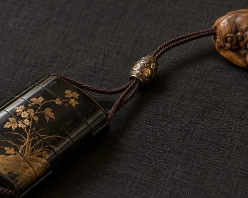 Illustratie van een metalen ojime-kraal met bloemendecoratie op koorden die een gelakte inro verbinden met een gesneden houten netsuke.